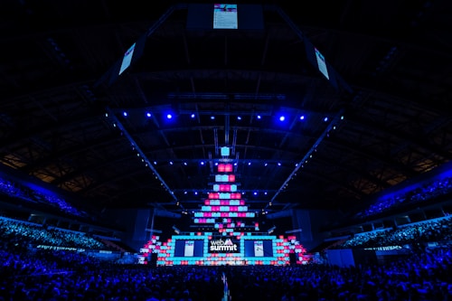 An expansive venue filled with a large audience, featuring a grand stage lit with vibrant blue and pink lights. The ceiling is decorated with a large display, and numerous screens showcase the event's branding. The atmosphere is dynamic and futuristic, reflecting a technologically advanced conference.