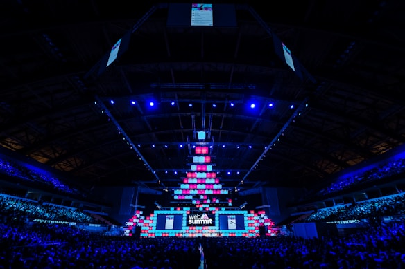 An expansive venue filled with a large audience, featuring a grand stage lit with vibrant blue and pink lights. The ceiling is decorated with a large display, and numerous screens showcase the event's branding. The atmosphere is dynamic and futuristic, reflecting a technologically advanced conference.