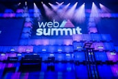 A large stage setup with an illuminated sign reading 'Web Summit' against a backdrop of stacked cube-like structures with purple and blue lighting. Several spotlights shine from above, and there is a seating arrangement that includes chairs and a table.