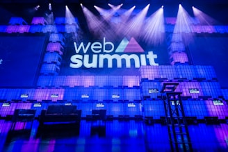 A large stage setup with an illuminated sign reading 'Web Summit' against a backdrop of stacked cube-like structures with purple and blue lighting. Several spotlights shine from above, and there is a seating arrangement that includes chairs and a table.