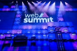 A large stage setup with an illuminated sign reading 'Web Summit' against a backdrop of stacked cube-like structures with purple and blue lighting. Several spotlights shine from above, and there is a seating arrangement that includes chairs and a table.