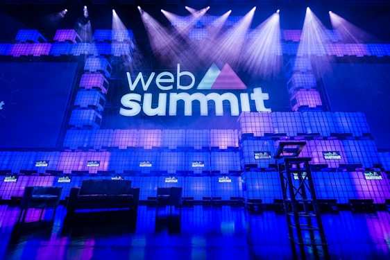 A large stage setup with an illuminated sign reading 'Web Summit' against a backdrop of stacked cube-like structures with purple and blue lighting. Several spotlights shine from above, and there is a seating arrangement that includes chairs and a table.