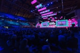 A large indoor tech conference with a vibrant stage setup featuring multiple screens displaying a speaker. The audience fills the arena, seated in rows, under a high ceiling with visible structural beams. The lighting emphasizes shades of blue and pink, contributing to a dynamic atmosphere.