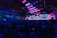 A large indoor tech conference with a vibrant stage setup featuring multiple screens displaying a speaker. The audience fills the arena, seated in rows, under a high ceiling with visible structural beams. The lighting emphasizes shades of blue and pink, contributing to a dynamic atmosphere.