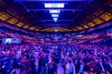 A large indoor venue filled with a massive crowd of people. The ceiling is high with a grid of lights illuminating the space, creating an atmosphere of anticipation. The seating areas are packed, and the central area is bustling with attendees engaged in conversation.