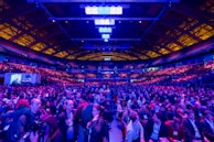 A large indoor venue filled with a massive crowd of people. The ceiling is high with a grid of lights illuminating the space, creating an atmosphere of anticipation. The seating areas are packed, and the central area is bustling with attendees engaged in conversation.