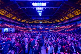 A large indoor venue filled with a massive crowd of people. The ceiling is high with a grid of lights illuminating the space, creating an atmosphere of anticipation. The seating areas are packed, and the central area is bustling with attendees engaged in conversation.