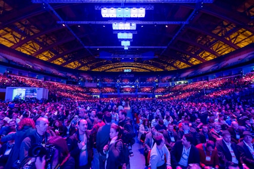 A large indoor venue filled with a massive crowd of people. The ceiling is high with a grid of lights illuminating the space, creating an atmosphere of anticipation. The seating areas are packed, and the central area is bustling with attendees engaged in conversation.