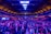 A large indoor venue filled with a massive crowd of people. The ceiling is high with a grid of lights illuminating the space, creating an atmosphere of anticipation. The seating areas are packed, and the central area is bustling with attendees engaged in conversation.