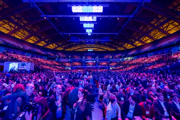 A large indoor venue filled with a massive crowd of people. The ceiling is high with a grid of lights illuminating the space, creating an atmosphere of anticipation. The seating areas are packed, and the central area is bustling with attendees engaged in conversation.