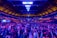 A large indoor venue filled with a massive crowd of people. The ceiling is high with a grid of lights illuminating the space, creating an atmosphere of anticipation. The seating areas are packed, and the central area is bustling with attendees engaged in conversation.