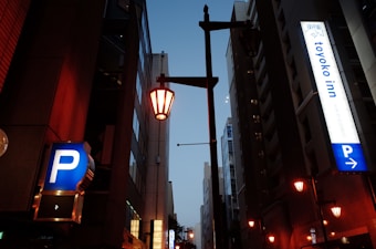 A nighttime urban scene featuring tall buildings on either side, illuminated street lamps, and signs for parking and a hotel. The signs include bright blue and white colors, with one sign displaying 'toyoko inn'. The street is dimly lit with an ambient glow from the lamps.