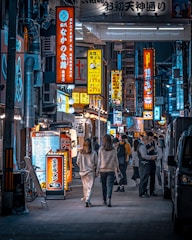 A bustling urban street scene features a vibrant display of illuminated signs in various shapes, sizes, and colors, mainly showcasing Japanese characters. Pedestrians, including two individuals walking side by side and others engaged in conversation, populate the sidewalk. The scene evokes the lively and energetic atmosphere of a city at night.