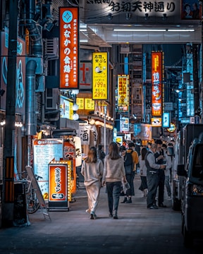 A bustling urban street scene features a vibrant display of illuminated signs in various shapes, sizes, and colors, mainly showcasing Japanese characters. Pedestrians, including two individuals walking side by side and others engaged in conversation, populate the sidewalk. The scene evokes the lively and energetic atmosphere of a city at night.