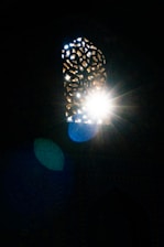 A decorative window with intricate patterns allows sunlight to stream through, creating a starburst effect. The light creates surrounding lens flares, casting hues of blue and green against a dimly lit wall.