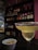 A cocktail glass filled with a light-colored drink rimmed with salt is placed on a bar counter. In the foreground, there is a small bowl containing pretzels. The background features shelves stocked with various bottles of liquor, against a backdrop of indirect lighting that creates a cozy atmosphere.