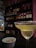 A cocktail glass filled with a light-colored drink rimmed with salt is placed on a bar counter. In the foreground, there is a small bowl containing pretzels. The background features shelves stocked with various bottles of liquor, against a backdrop of indirect lighting that creates a cozy atmosphere.