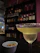 A cocktail glass filled with a light-colored drink rimmed with salt is placed on a bar counter. In the foreground, there is a small bowl containing pretzels. The background features shelves stocked with various bottles of liquor, against a backdrop of indirect lighting that creates a cozy atmosphere.