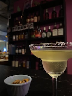 A cocktail glass filled with a light-colored drink rimmed with salt is placed on a bar counter. In the foreground, there is a small bowl containing pretzels. The background features shelves stocked with various bottles of liquor, against a backdrop of indirect lighting that creates a cozy atmosphere.