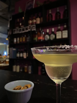 A cocktail glass filled with a light-colored drink rimmed with salt is placed on a bar counter. In the foreground, there is a small bowl containing pretzels. The background features shelves stocked with various bottles of liquor, against a backdrop of indirect lighting that creates a cozy atmosphere.