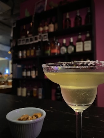 A cocktail glass filled with a light-colored drink rimmed with salt is placed on a bar counter. In the foreground, there is a small bowl containing pretzels. The background features shelves stocked with various bottles of liquor, against a backdrop of indirect lighting that creates a cozy atmosphere.