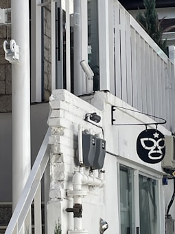 A building exterior features white and gray tones with visible pipes and meters attached to the wall. A black sign with a stylized mask hangs on the wall. The structure includes modern design elements like glass windows and white panel railings.