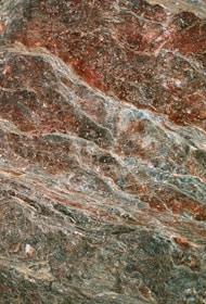 The image displays a close-up view of a textured rock surface with swirling patterns and layers. The rock features rich combinations of earthy red and grey tones, exhibiting a natural, rugged texture.
