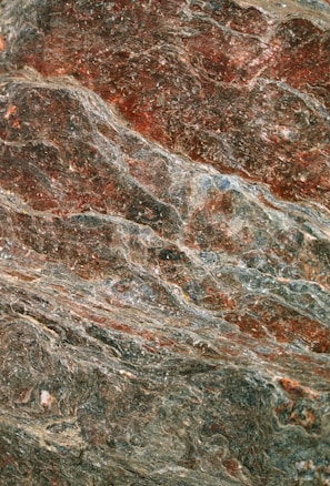The image displays a close-up view of a textured rock surface with swirling patterns and layers. The rock features rich combinations of earthy red and grey tones, exhibiting a natural, rugged texture.