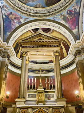 An ornate altar is beautifully adorned with intricate carvings, featuring tall columns and a central crucifix. The surrounding architecture includes detailed frescoes depicting religious scenes on the ceiling. Warm lighting highlights the textures and colors of the intricate designs.
