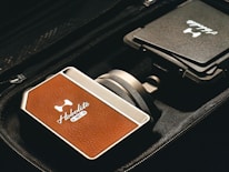 A close-up view of a device with a brown leather-like texture, branded 'Hobolite' and labeled with 'NFC' in a sleek, metallic case. The device is positioned next to a folded item with another 'Hobolite' label, all placed within a black fabric bag or case.