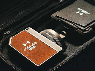 A close-up view of a device with a brown leather-like texture, branded 'Hobolite' and labeled with 'NFC' in a sleek, metallic case. The device is positioned next to a folded item with another 'Hobolite' label, all placed within a black fabric bag or case.