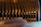 A modern wine display featuring a series of large bottles suspended in an artistic arrangement against a dark background. Vertical wooden slats are evenly spaced, creating a wave-like pattern. A curved, plush bench sits in the foreground, adding a luxurious touch to the setting.