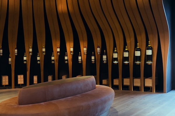 A modern wine display featuring a series of large bottles suspended in an artistic arrangement against a dark background. Vertical wooden slats are evenly spaced, creating a wave-like pattern. A curved, plush bench sits in the foreground, adding a luxurious touch to the setting.