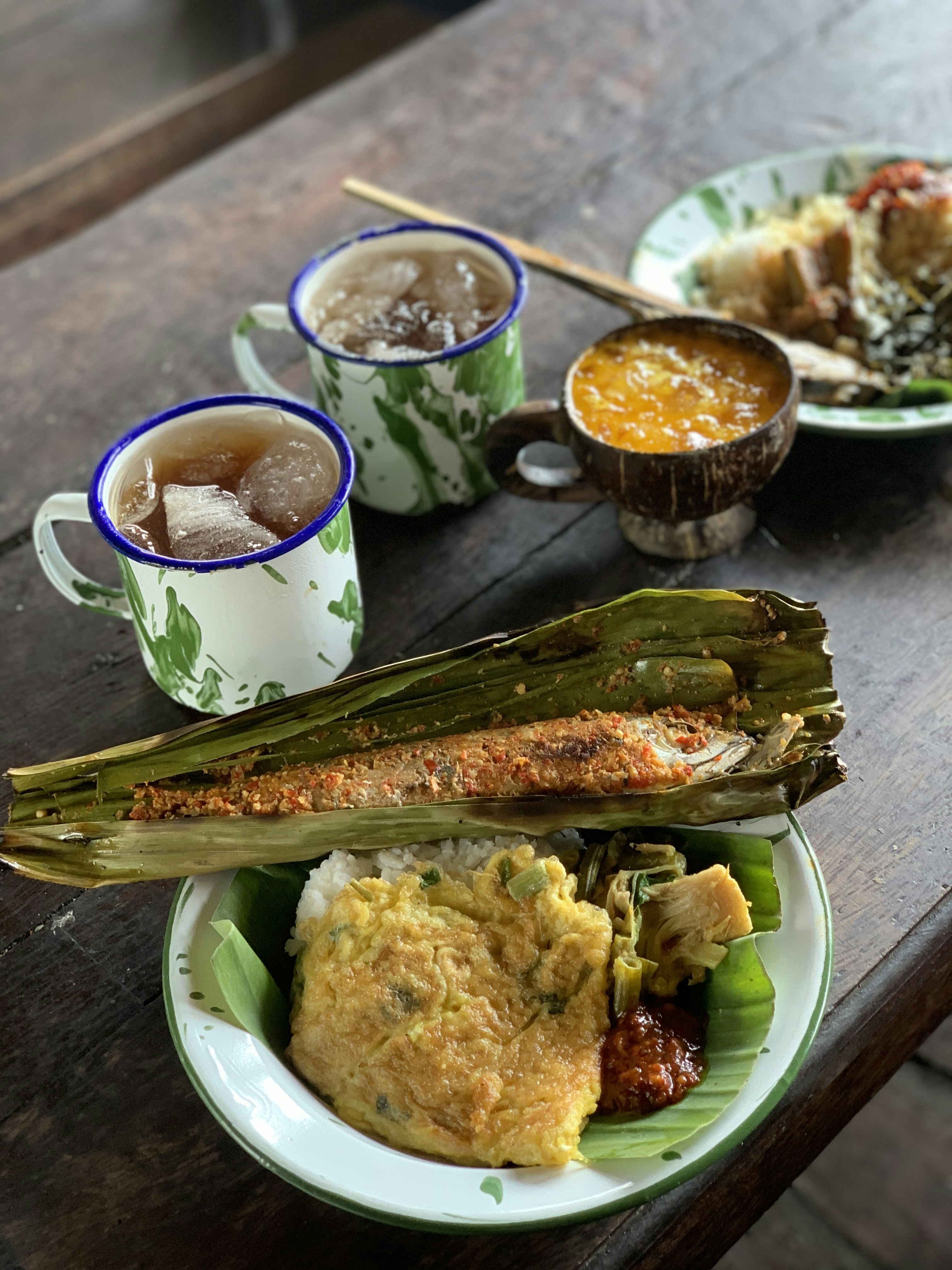 raditional Indonesian meal served on an enamel plate with rice, grilled fish in banana leaf (pepes ikan), omelette, vegetables, and sambal, accompanied by iced tea in vintage enamel mugs and a dessert in a coconut shell bowl.