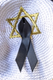 A black ribbon is placed on a white woven surface with a gold-colored embroidered Star of David.
