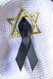 A black ribbon is placed on a white woven surface with a gold-colored embroidered Star of David.
