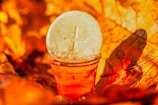 A white communion wafer with a cross imprint stands propped against a small glass filled with a red liquid, possibly wine or juice. The background is filled with orange and yellow hues, suggesting autumn leaves, creating a warm and vibrant atmosphere.