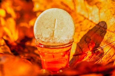 A white communion wafer with a cross imprint stands propped against a small glass filled with a red liquid, possibly wine or juice. The background is filled with orange and yellow hues, suggesting autumn leaves, creating a warm and vibrant atmosphere.