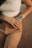 Close-up of a person wearing a white crop top with 'Angel Baby' written on it, tan pants, and wrist accessories including a digital watch and multiple bracelets. The hands are clasped together, displaying manicured nails against a textured background.