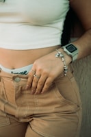 A woman wearing a white crop top and beige pants partially visible, showcasing a smartwatch on her wrist and a silver bracelet. Her hand rests on her hip, adorned with a ring, against a textured background.