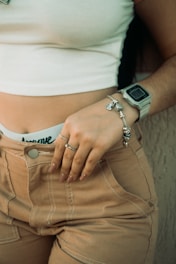A woman wearing a white crop top and beige pants partially visible, showcasing a smartwatch on her wrist and a silver bracelet. Her hand rests on her hip, adorned with a ring, against a textured background.