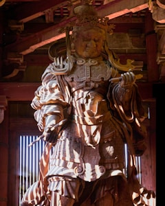 A detailed wooden statue of an armored warrior is positioned in a traditional wooden structure. The intricate armor and ornate crown suggest historical or cultural significance. Light and shadows play across the surface, highlighting the handcrafted details and expressions on the statue's face.