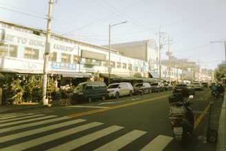 A street scene featuring a market on the left with various stalls, some selling plants. Cars are parked along the road, and there is a visible zebra crossing in the foreground leading towards the market. The sky is clear and there are visible power lines.