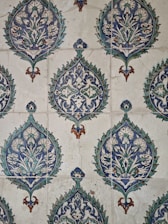 The image showcases a detailed pattern of decorative ceramic tiles. Each tile features intricate floral and geometric designs primarily in shades of blue, green, and white, with accents of brown. The repeating pattern consists of a central floral motif encased within a teardrop shape adorned with foliage and symmetrical designs extending outward.