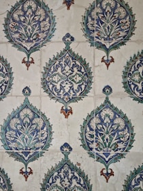 The image showcases a detailed pattern of decorative ceramic tiles. Each tile features intricate floral and geometric designs primarily in shades of blue, green, and white, with accents of brown. The repeating pattern consists of a central floral motif encased within a teardrop shape adorned with foliage and symmetrical designs extending outward.