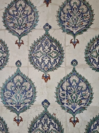 The image showcases a detailed pattern of decorative ceramic tiles. Each tile features intricate floral and geometric designs primarily in shades of blue, green, and white, with accents of brown. The repeating pattern consists of a central floral motif encased within a teardrop shape adorned with foliage and symmetrical designs extending outward.