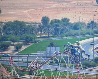 A dirt bike rider is performing a stunt, leaping over a rollercoaster track in an arid, desert-like environment. Greenery and palm trees fringe the landscape, with highways and a few cars visible in the background.