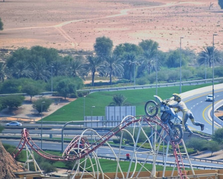 A dirt bike rider is performing a stunt, leaping over a rollercoaster track in an arid, desert-like environment. Greenery and palm trees fringe the landscape, with highways and a few cars visible in the background.