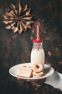 A hand holds a white plate containing cookies and a bottle of milk. The cookies include jam-filled biscuits and rectangular ones with the word 'NICE' embossed on them. The milk is in a retro glass bottle with a red cap and a red and white striped straw. In the background, a decorative paper star is visible against a textured, dark surface.