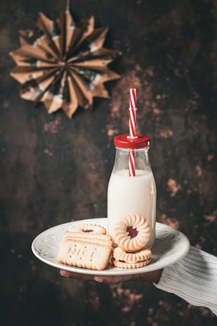 A hand holds a white plate containing cookies and a bottle of milk. The cookies include jam-filled biscuits and rectangular ones with the word 'NICE' embossed on them. The milk is in a retro glass bottle with a red cap and a red and white striped straw. In the background, a decorative paper star is visible against a textured, dark surface.
